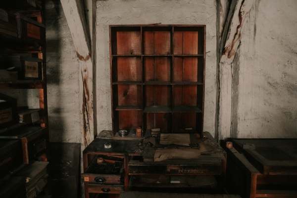 Rotbrauner Fächerschrank an der geweißten Wand, davor ein Tisch mit Tintenfass und Papieren