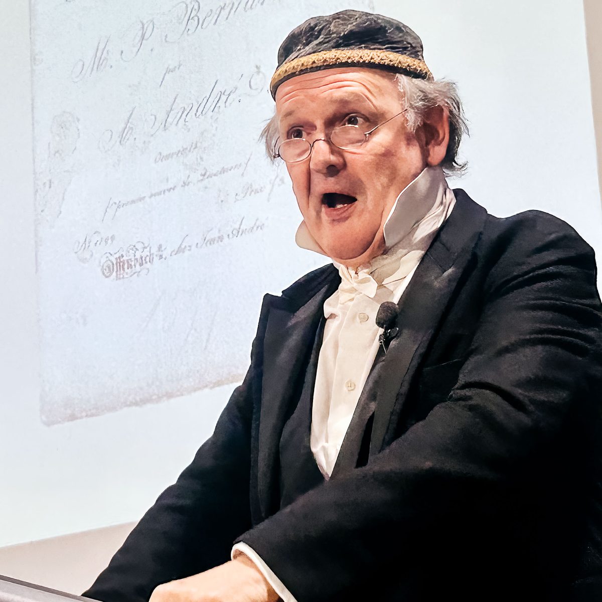Michael Quast als Johann Anton André: dunkler Rock, weiße Halsbinde und besticktes Käppchen, im Sprechen vor der projizierten Titelseite eines André-Drucks.