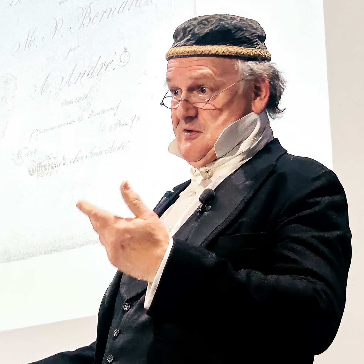Michael Quast als Johann Anton André im Halbprofil am Rednerpult, den Zeigefinger erhoben.