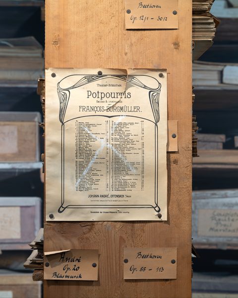Verlagsverzeichnis „Theater-Bibliothek. Potpourris faciles & instructifs par François Burgmüller“ des Verlags Johann André am Pfosten eines Notenregals, darüber und darunter handbeschriftete Fachschilder.