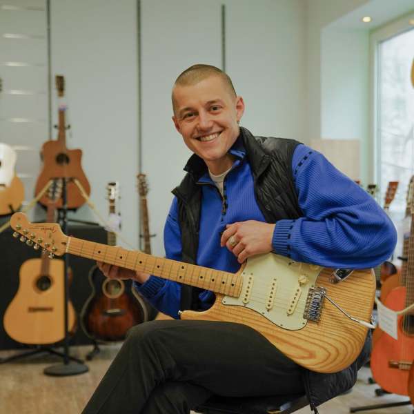 Moritz André sitzt lachend mit einer E-Gitarre im Musikhaus André, hinter ihm eine Wand voller Gitarren